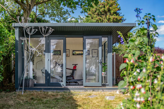 Modern grey garden room with floor-to-ceiling glazing and double doors, set among mature garden planting and used as a home gym.